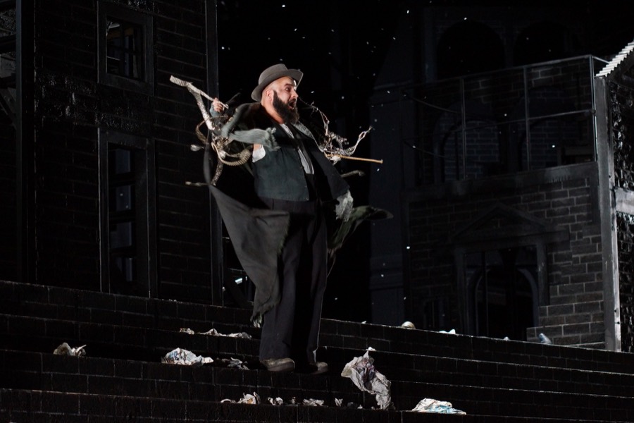 Ralf Rachbauer als Mastro Trabucco in »La Forza del Destino« an der Oper Köln: auf einer Treppe stehend, Papier am Boden verstreut